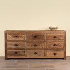A rustic 9-drawer wooden dresser made of warm medium-brown wood with dark metal round pulls, set against a plain beige wall and brick floor.