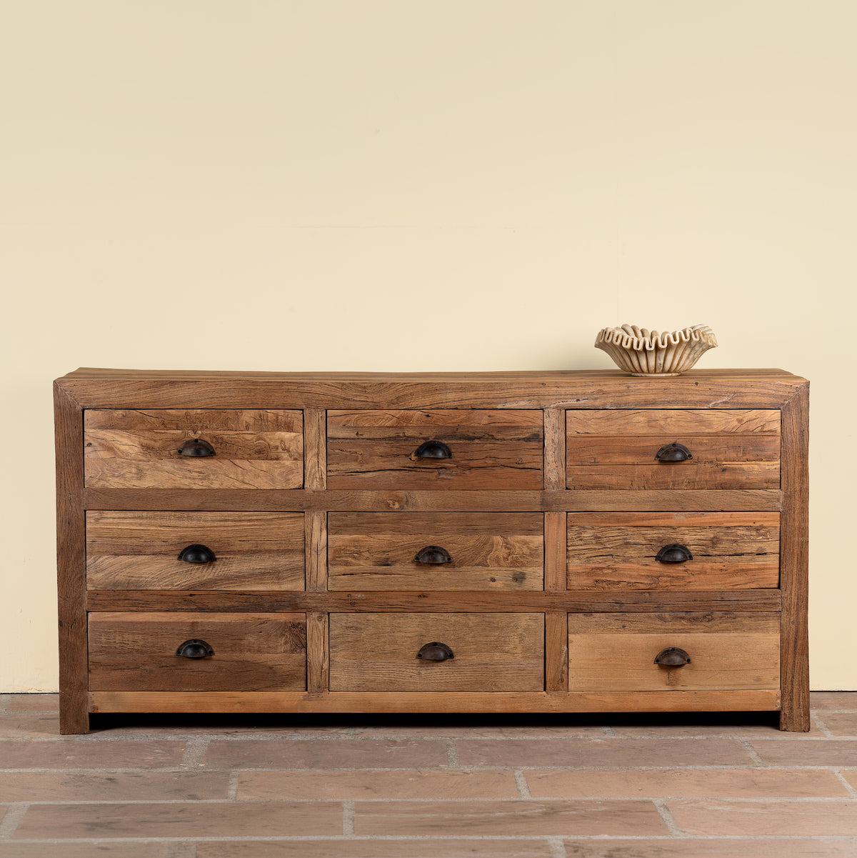 A rustic 9-drawer wooden dresser made of warm medium-brown wood with dark metal round pulls, set against a plain beige wall and brick floor.