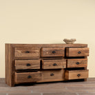 A rustic wooden 9-drawer dresser with a weathered brown finish and dark metal knobs, set against a plain beige wall and brown tiled floor.