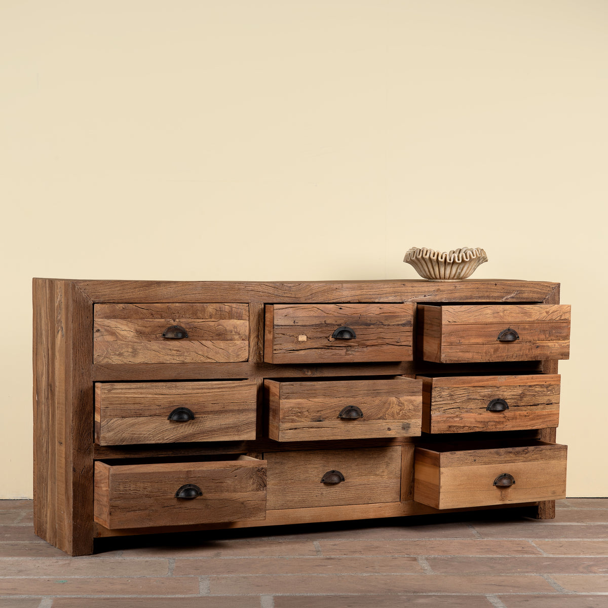 A rustic wooden 9-drawer dresser with a weathered brown finish and dark metal knobs, set against a plain beige wall and brown tiled floor.
