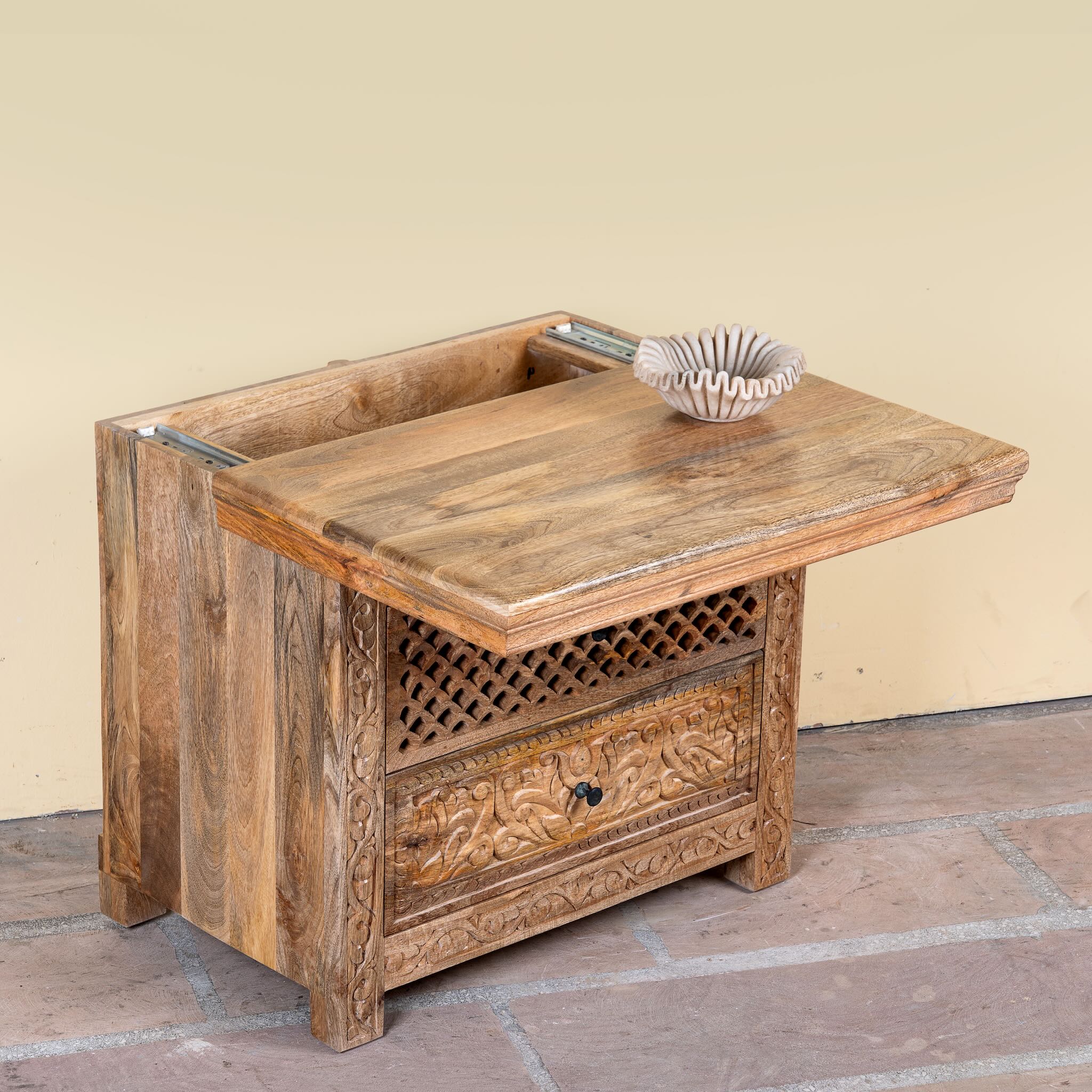 Side angle image showing open lid of a hand carved mango wood nightstand with two drawers and hidden compartment in rustic artisan style.
