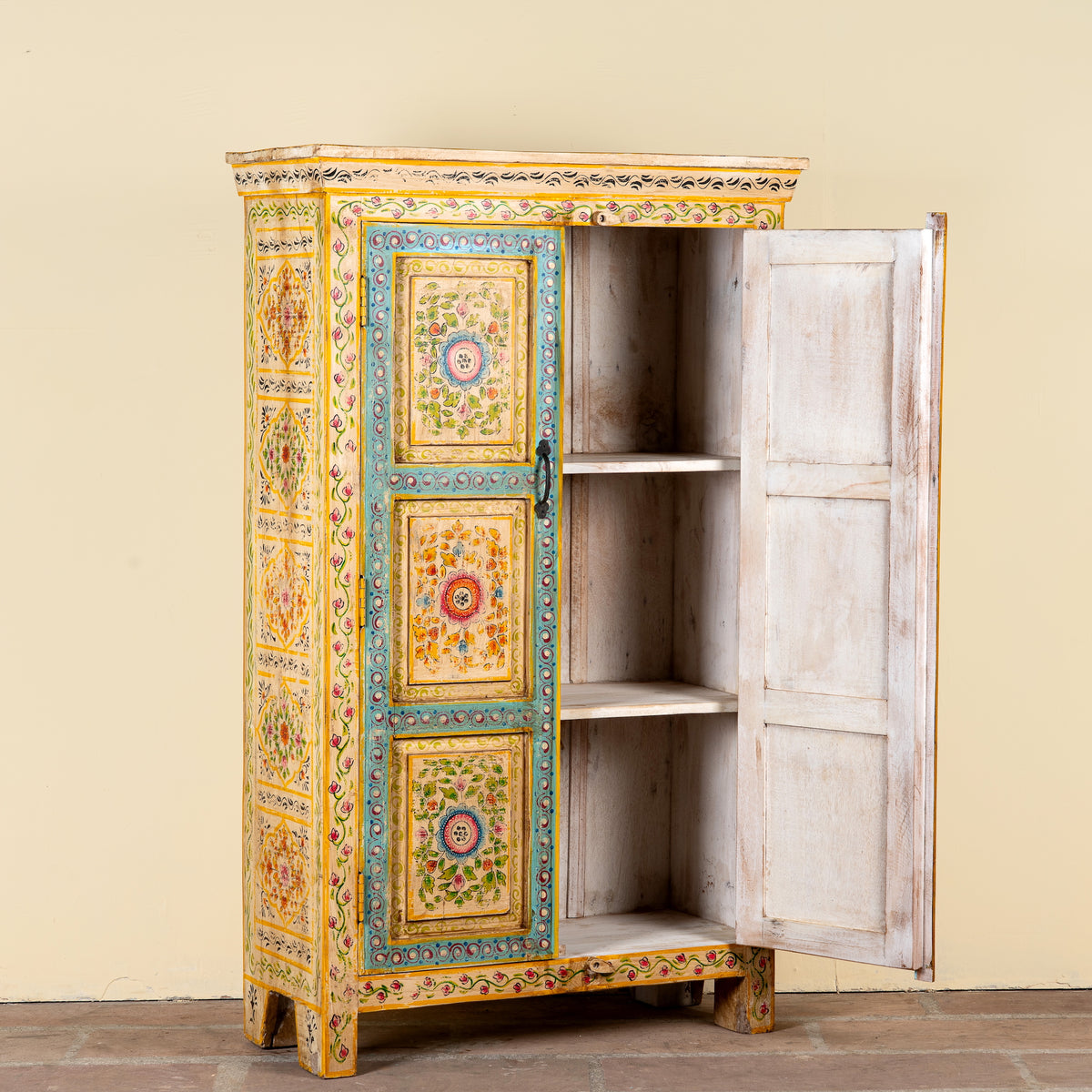 An ornate hand-painted wooden cabinet with a distressed white-painted right door open to reveal three shelves, set against a plain beige wall.