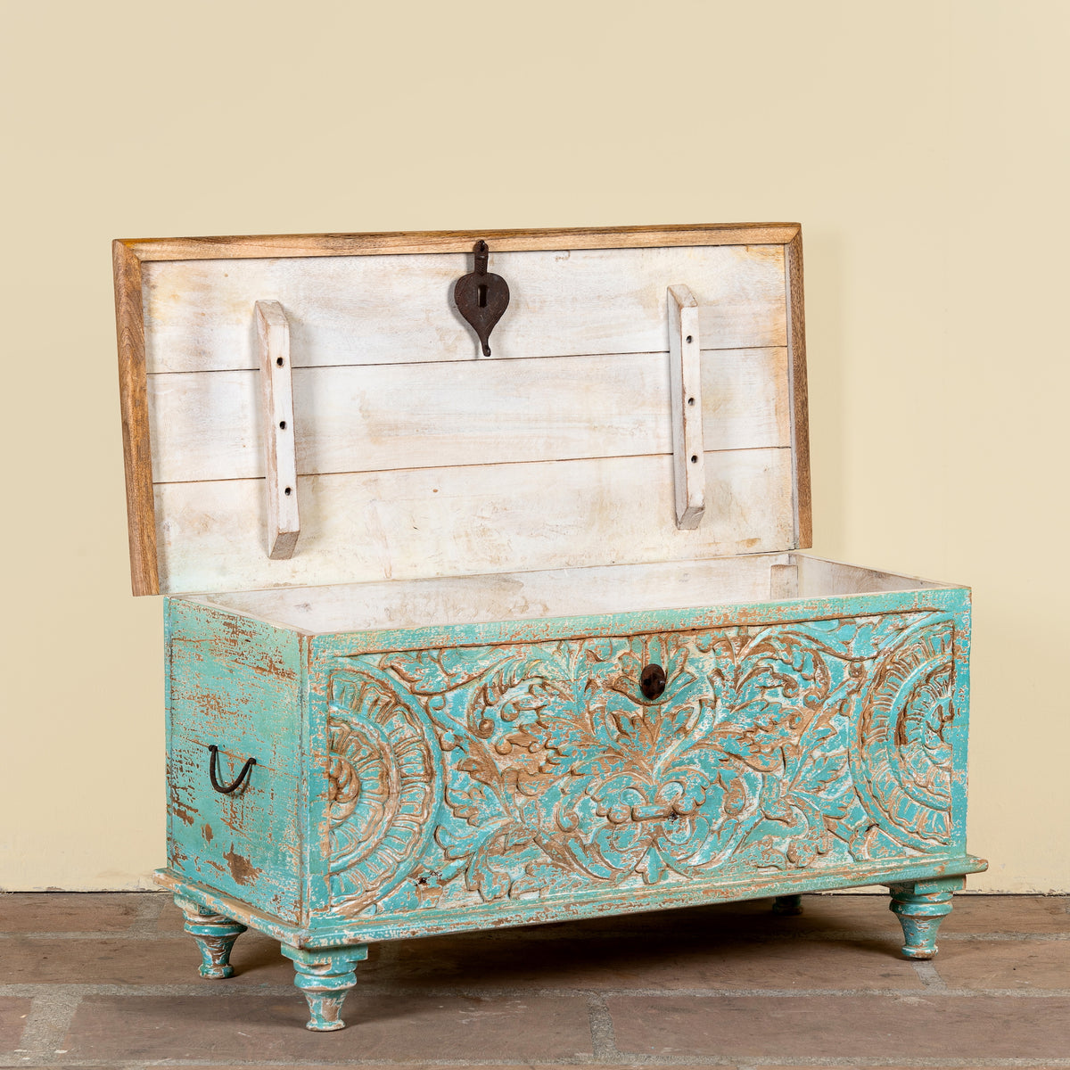 A distressed turquoise and white carved wooden chest with an open hinged lid, shown against a plain beige wall and rustic wood floor.