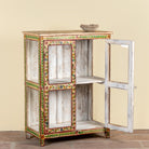 A rustic hand-painted wooden cabinet with a distressed white finish and colorful floral and geometric borders in green, red, and gold. The cabinet features two open side compartments and a distressed white door on the right, shown against a plain beige wall.
