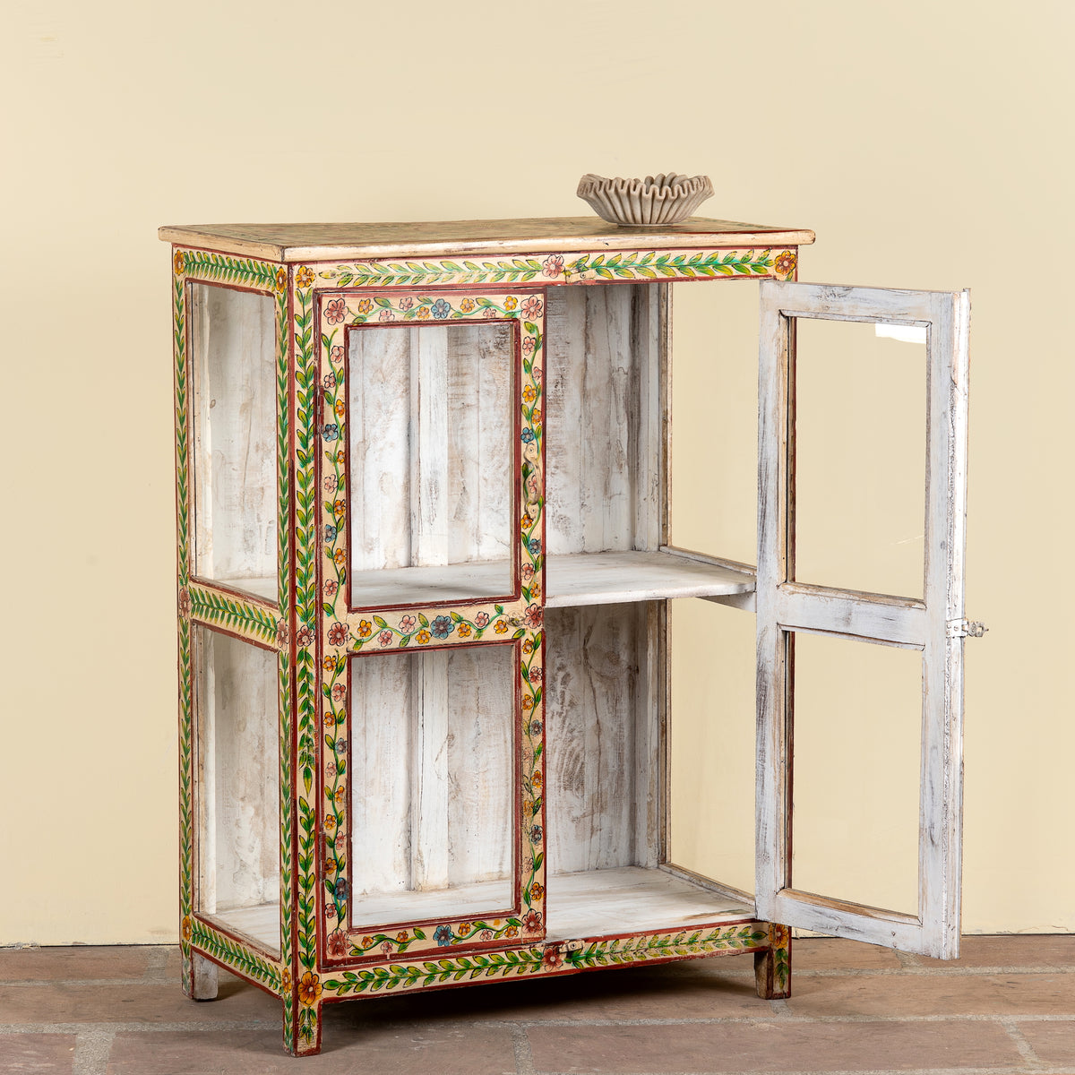 A rustic hand-painted wooden cabinet with a distressed white finish and colorful floral and geometric borders in green, red, and gold. The cabinet features two open side compartments and a distressed white door on the right, shown against a plain beige wall.