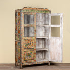 A tall, hand-painted wooden cabinet with a distressed white finish on the doors and shelves, decorated with colorful traditional Indian motifs including peacocks and elephants. The cabinet stands open against a plain beige wall.