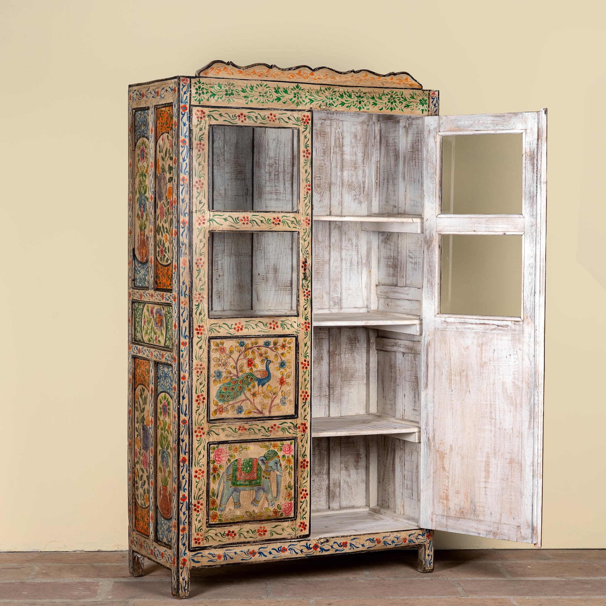 A tall, hand-painted wooden cabinet with a distressed white finish on the doors and shelves, decorated with colorful traditional Indian motifs including peacocks and elephants. The cabinet stands open against a plain beige wall.