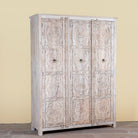 Side angle image of a large hand carved mango wood wardrobe with floral carvings, whitewashed finish, four doors, and drawer storage.