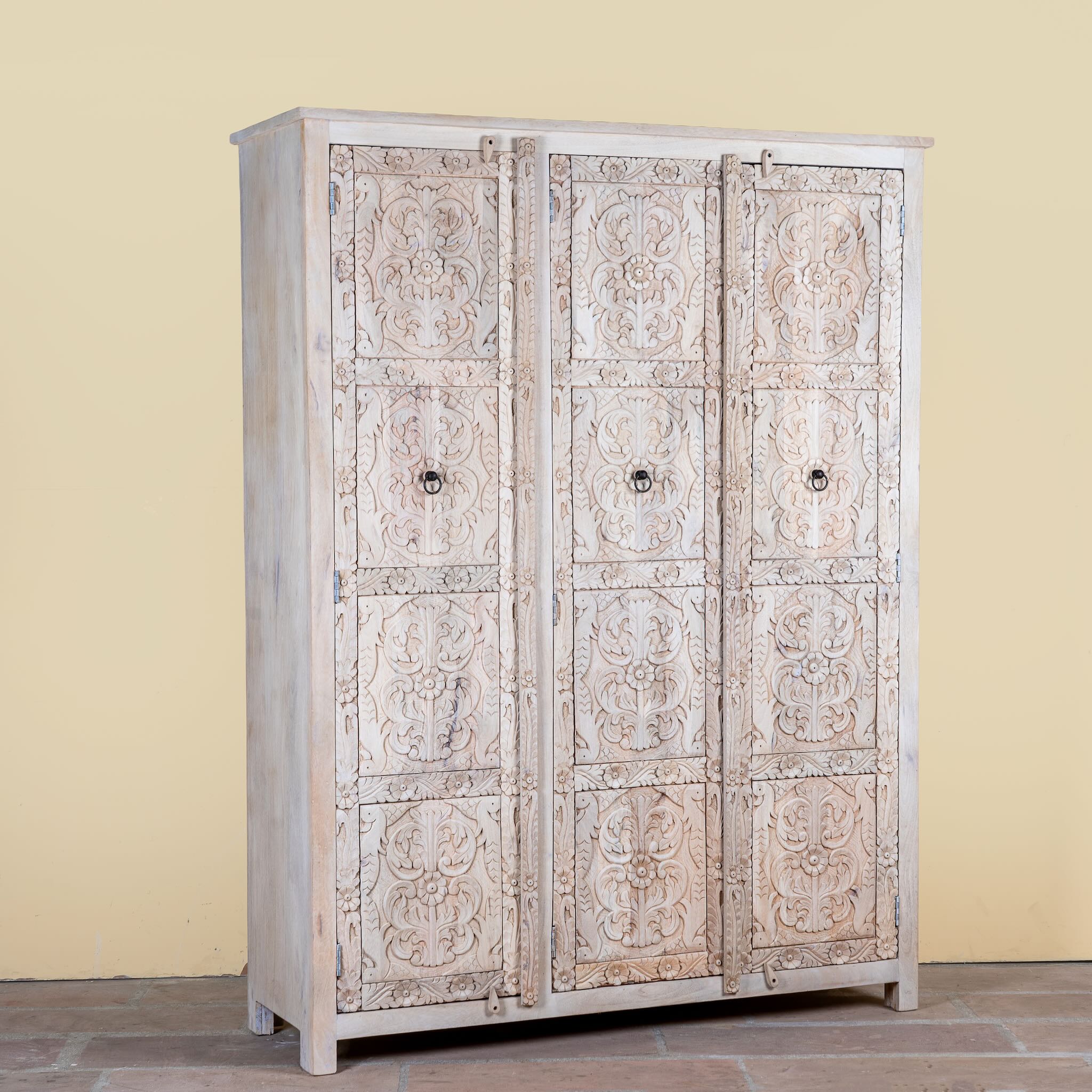 Side angle image of a large hand carved mango wood wardrobe with floral carvings, whitewashed finish, four doors, and drawer storage.