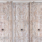 Close up image of a large hand carved mango wood wardrobe with floral carvings, whitewashed finish, four doors, and drawer storage.