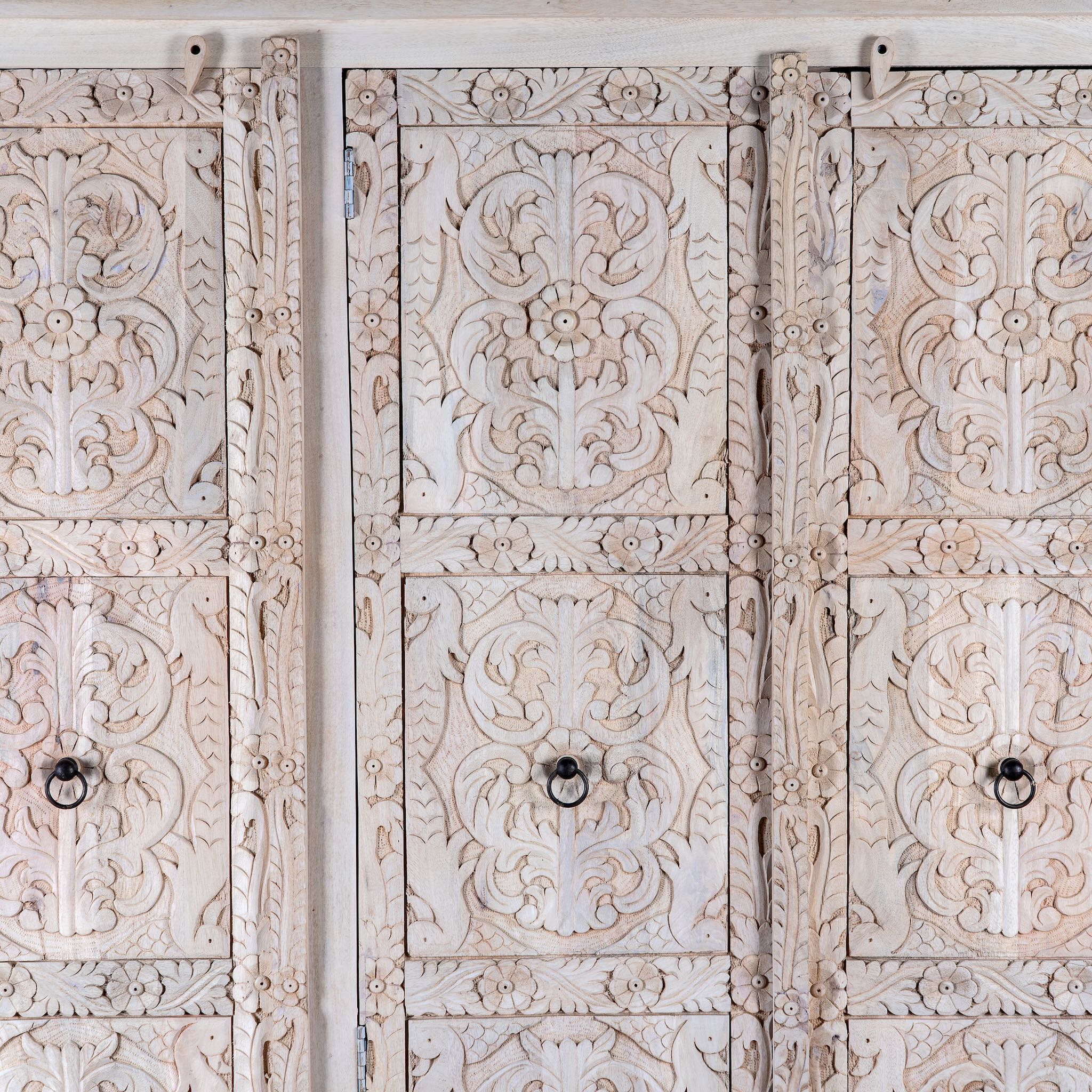 Close up image of a large hand carved mango wood wardrobe with floral carvings, whitewashed finish, four doors, and drawer storage.