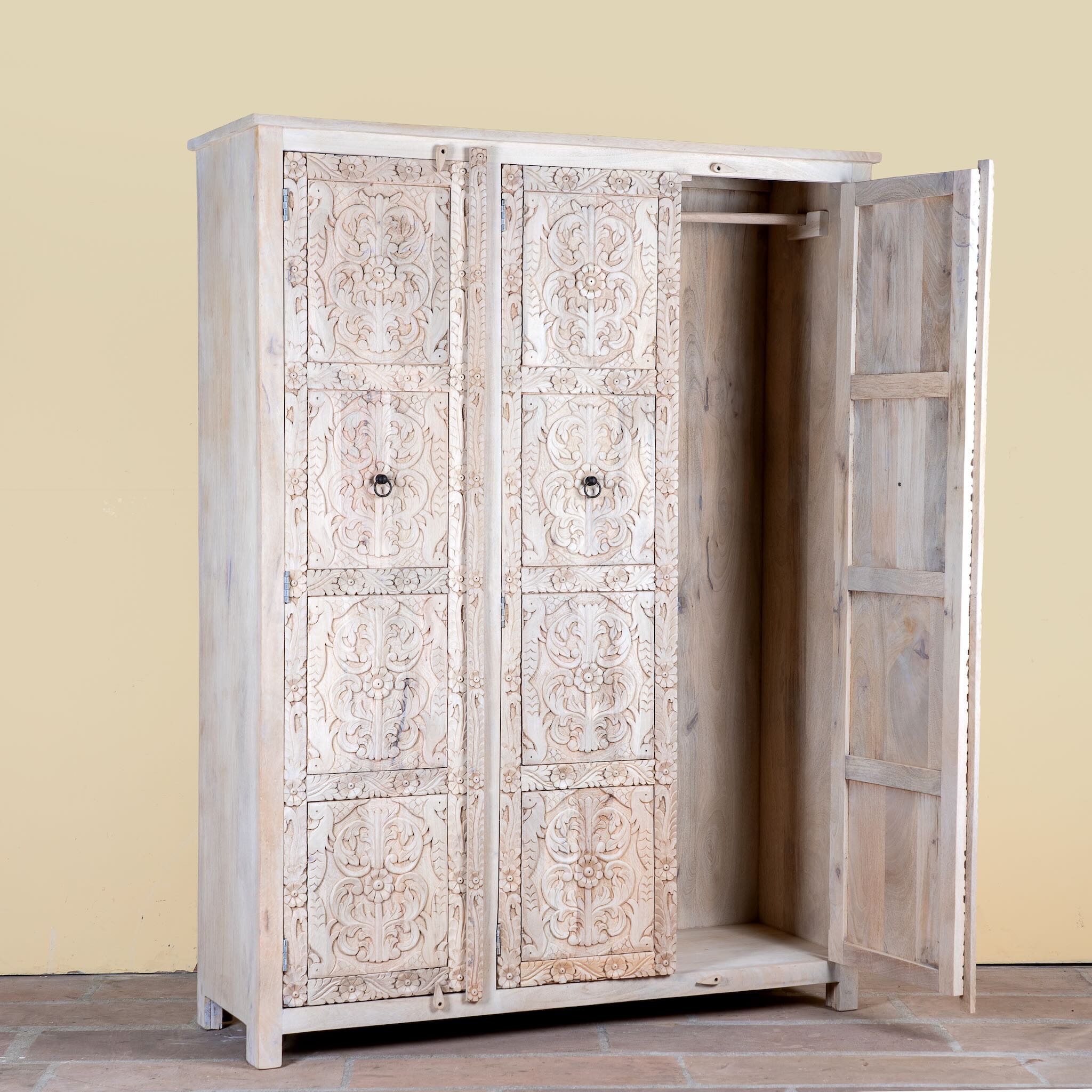 Side angle image with open door of a large hand carved mango wood wardrobe with floral carvings, whitewashed finish, four doors, and drawer storage.