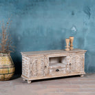 Side angle image of a carved mango wood lowboard with floral doors, crackled vintage finish, drawer, and open shelf, 150 × 40 × 64 cm.