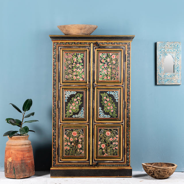 A hand-painted wooden wardrobe with floral patterns in vibrant colors on a dark background, positioned against a blue wall with a decorative plate above it. There is also a wooden bowl on top of the wardrobe and a plant to the left.
