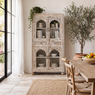 Lifestyle image of a hand-carved mango wood showcase with glass doors and drawers – carved wooden furniture in distressed turquoise and blue, combining artisan detail with vintage charm.