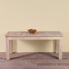 A light natural wood dining table with a slatted top and block legs, shown against a plain beige wall and brick floor with a small woven bowl on top.