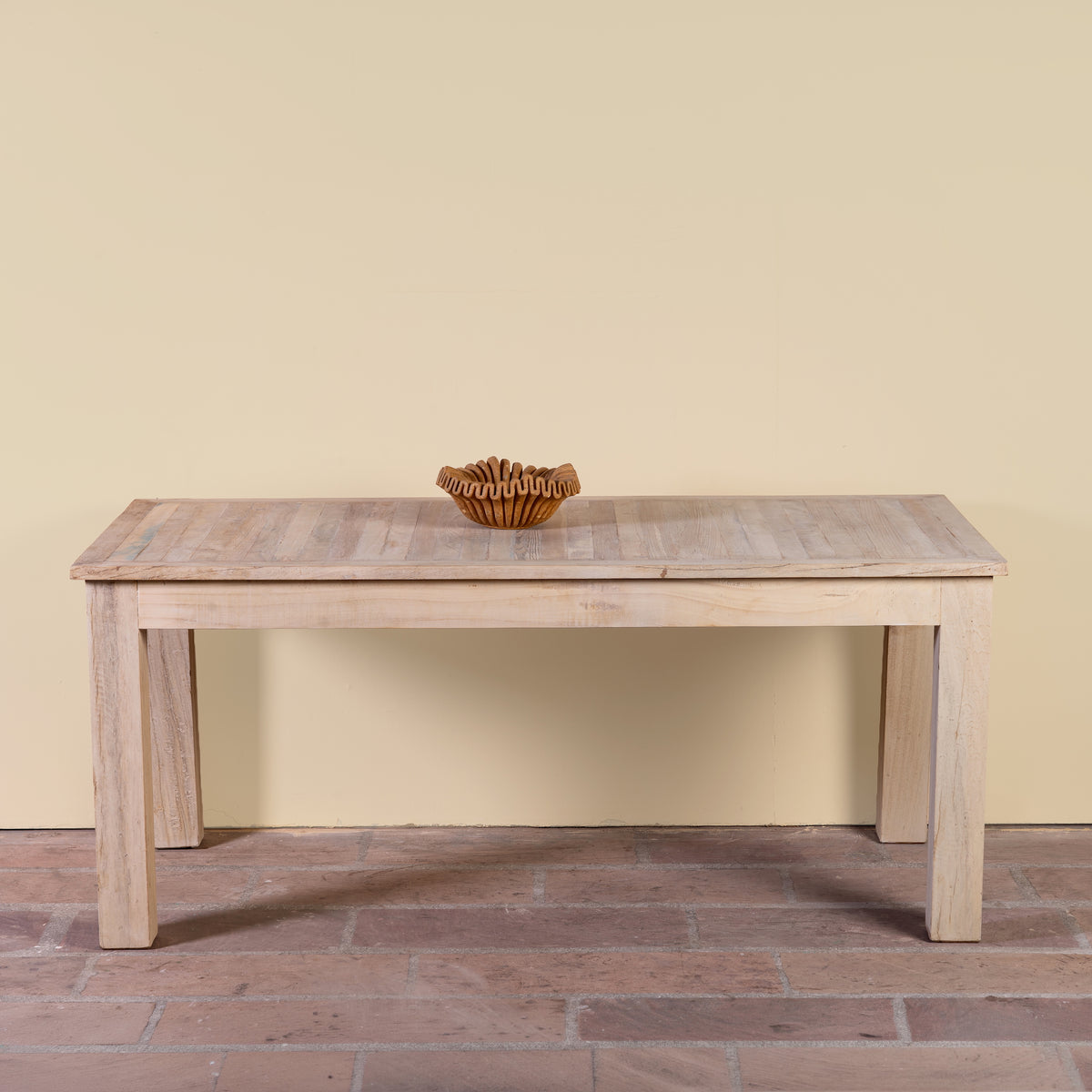 A light natural wood dining table with a slatted top and block legs, shown against a plain beige wall and brick floor with a small woven bowl on top.