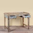 An ornate antique-style writing desk with a distressed beige wood finish, decorated with blue and gold floral inlays on the drawer fronts and aprons, and a matching inlaid top surface, set against a plain beige wall and stone floor.