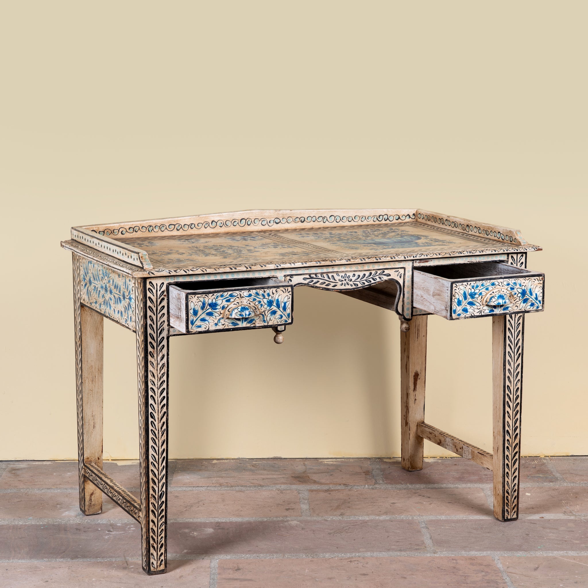 An ornate antique-style writing desk with a distressed beige wood finish, decorated with blue and gold floral inlays on the drawer fronts and aprons, and a matching inlaid top surface, set against a plain beige wall and stone floor.