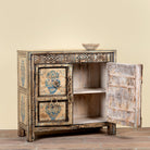 A rustic hand-painted wooden cabinet with a distressed finish, featuring blue floral motifs on the doors and ornate geometric carvings. The cabinet has one open door showing an interior shelf, and a small decorative bowl sits on top against a solid beige background.