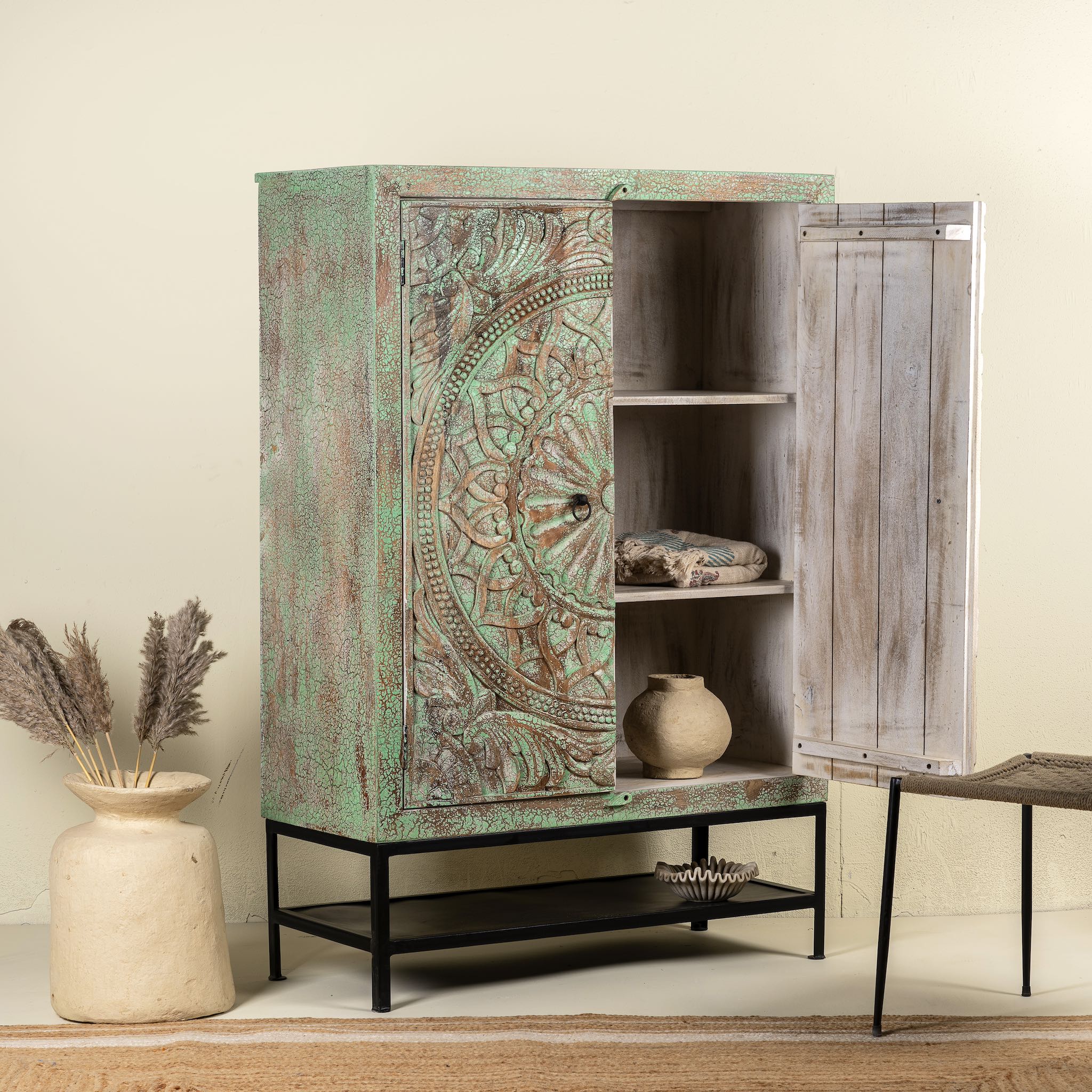 Open door image of a carved mango wood cabinet with floral hand-carving, distressed green finish, iron stand, 100 × 40 × 150 cm.