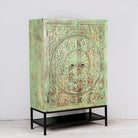 Aide angle image of a carved mango wood cabinet with floral hand-carving, distressed green finish, iron stand, 100 × 40 × 150 cm.