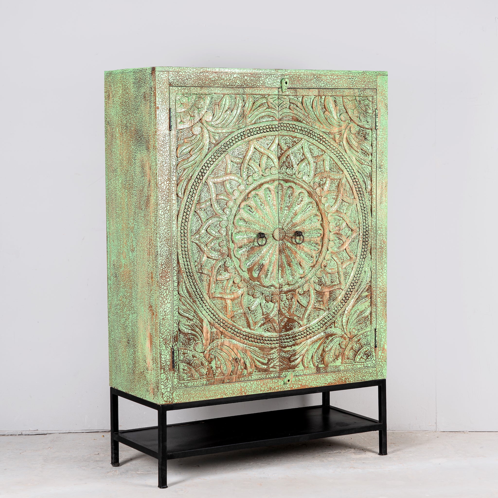 Aide angle image of a carved mango wood cabinet with floral hand-carving, distressed green finish, iron stand, 100 × 40 × 150 cm.