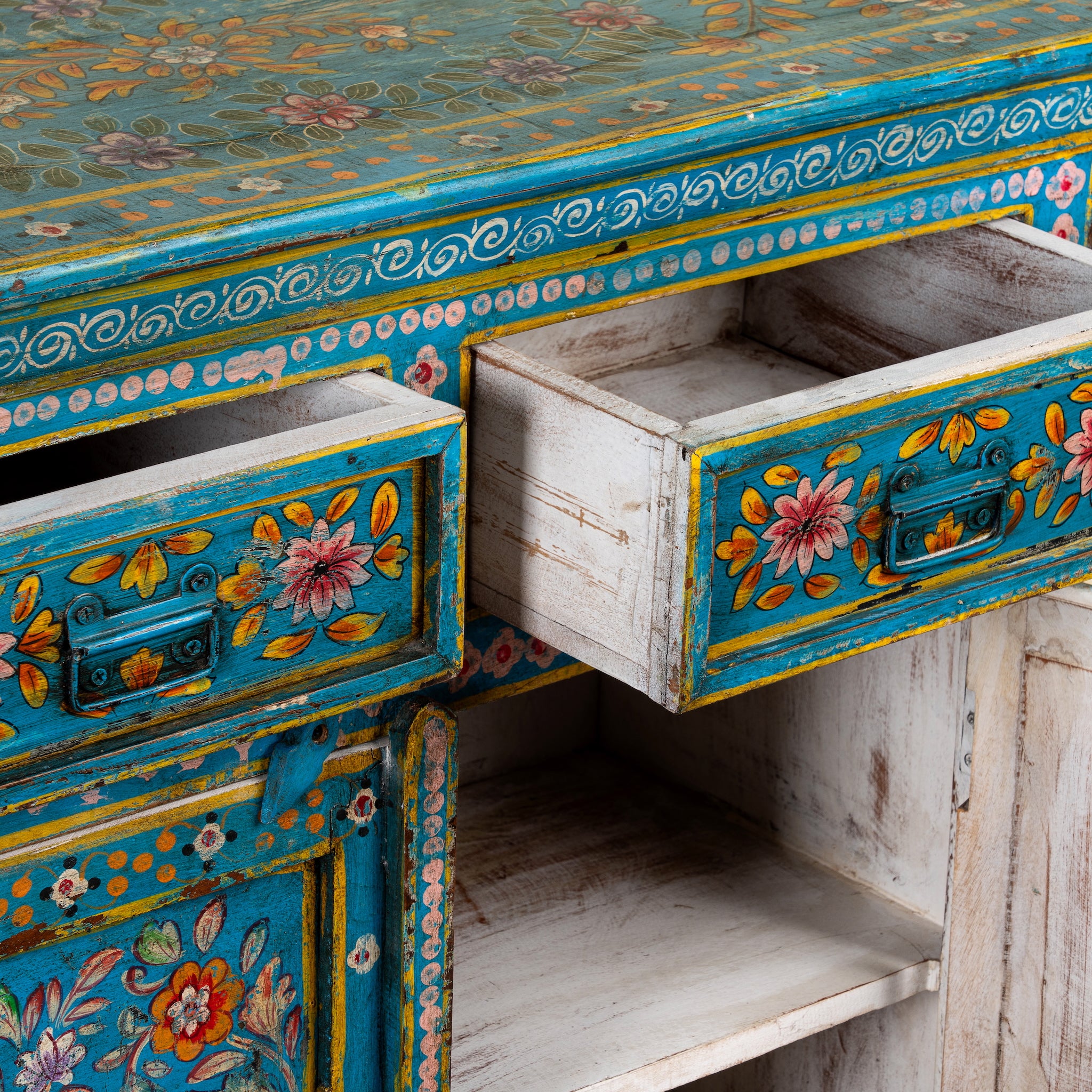 A close-up of a turquoise and white distressed wooden cabinet with colorful floral hand-painted details and two open drawers.