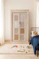 Lifestyle imagination of a whitewashed mango wood cabinet with hand-carved details, rattan inserts, and glass panels; versatile as bedroom furniture or wooden wardrobe; 192 × 110 × 40 cm.