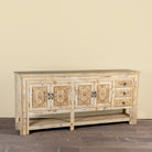 Side angle image of a Ranjit reclaimed wood vintage console table with cream distressed finish, carved doors, three drawers and open lower shelf.