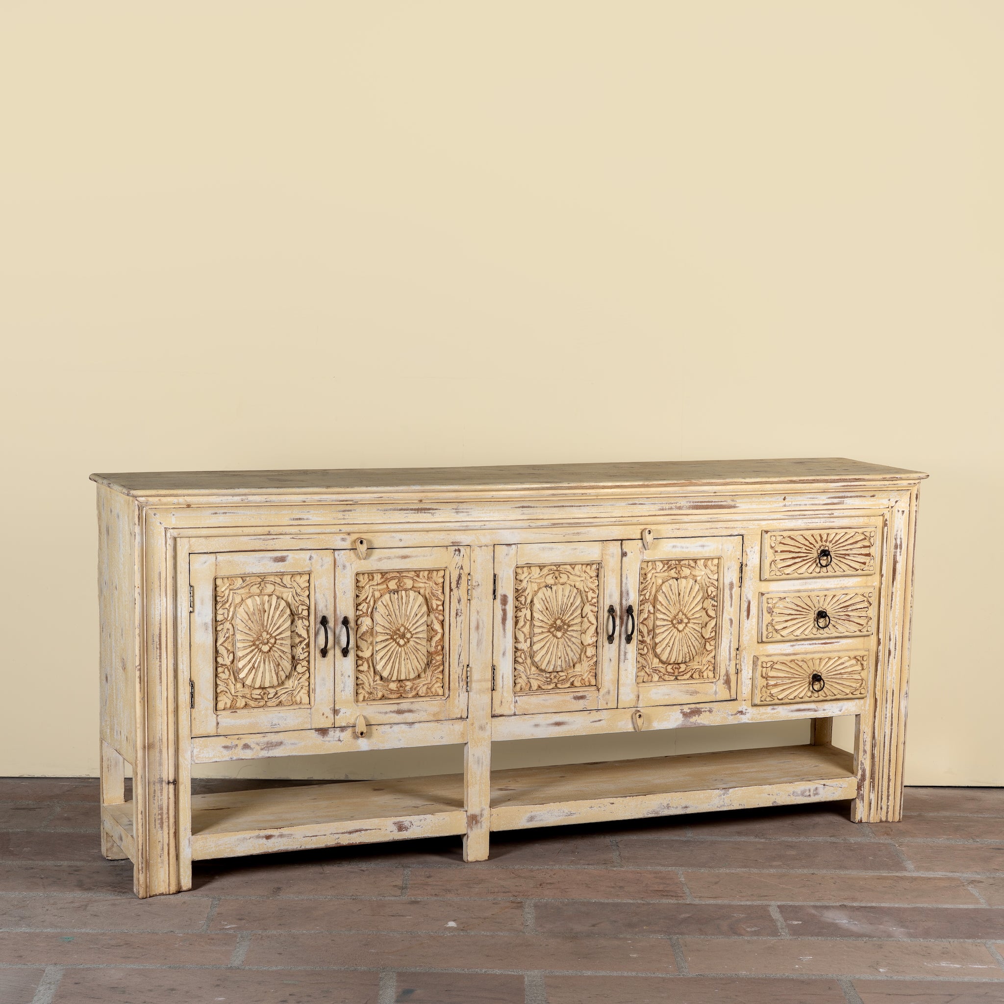 Side angle image of a Ranjit reclaimed wood vintage console table with cream distressed finish, carved doors, three drawers and open lower shelf.