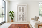White cabinet with decorative patterns in a living room setting with a plant and sofa.