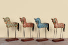 Four decorative hand-painted wooden horse statues with black tails and metal legs, standing on rectangular wooden bases against a plain beige wall.