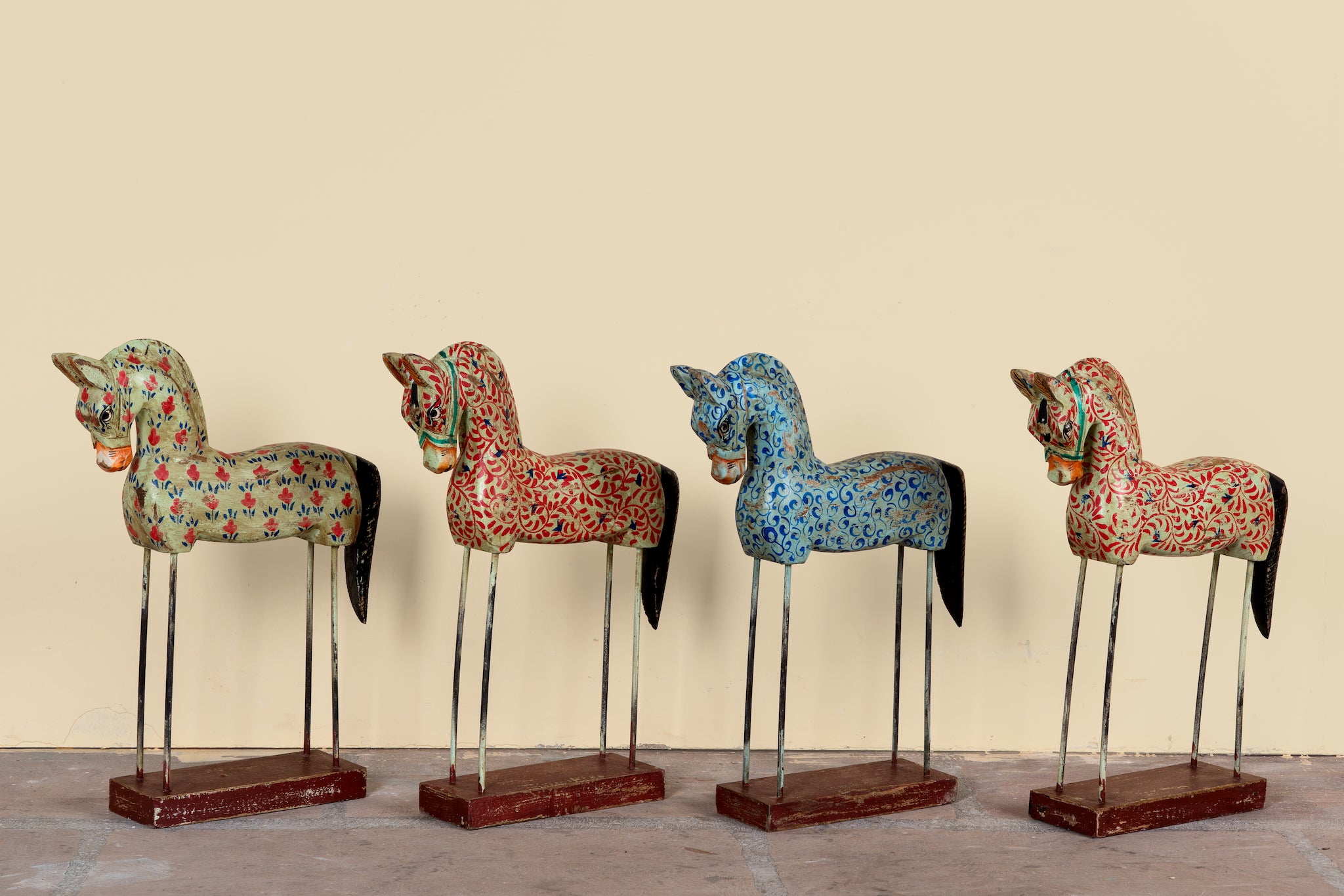 Four decorative hand-painted wooden horse statues with black tails and metal legs, standing on rectangular wooden bases against a plain beige wall.