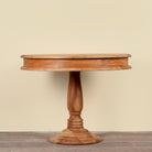A round wooden pedestal table with a warm medium-brown finish, featuring a turned central base and a hinged top, set against a plain beige wall and wooden floor.