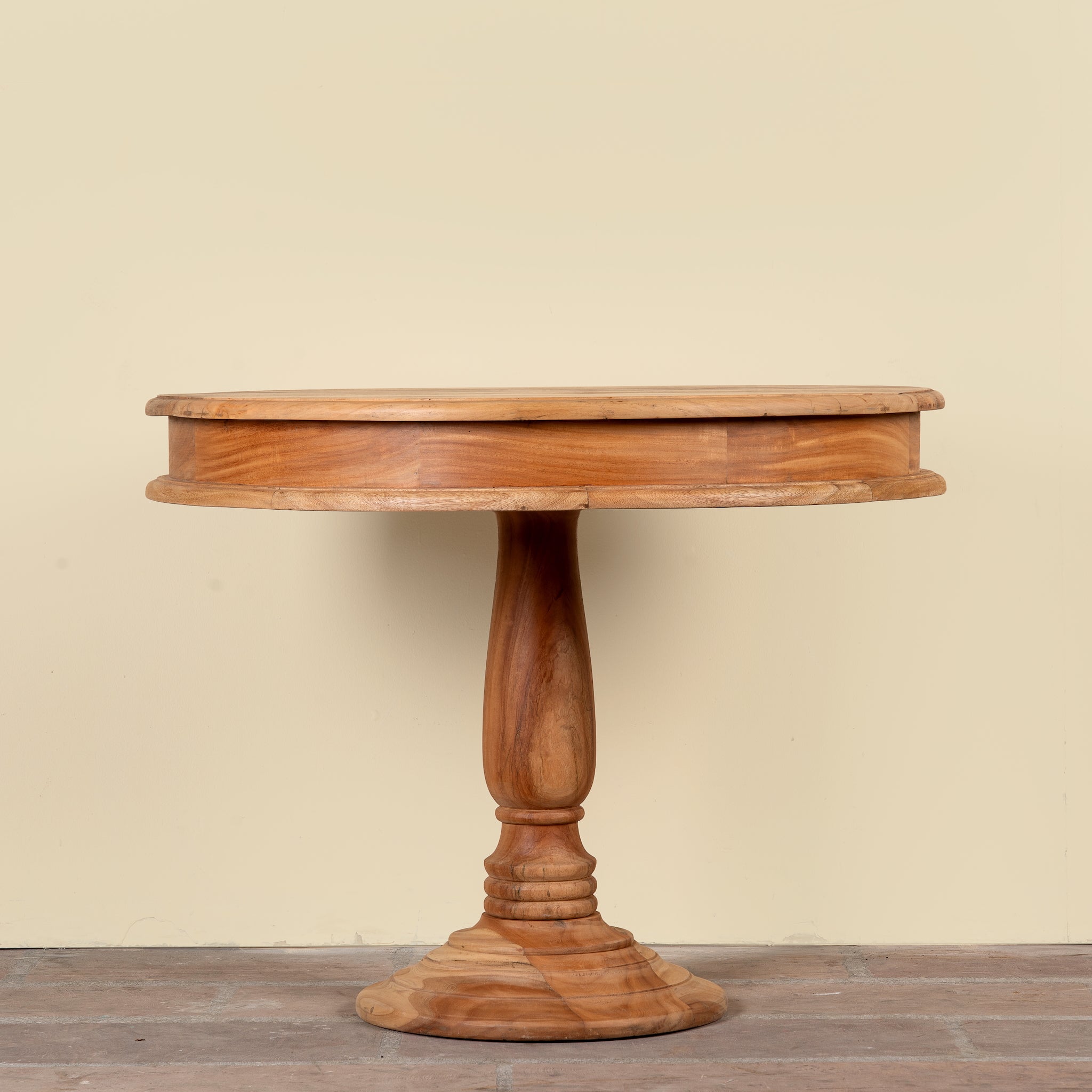 A round wooden pedestal table with a warm medium-brown finish, featuring a turned central base and a hinged top, set against a plain beige wall and wooden floor.