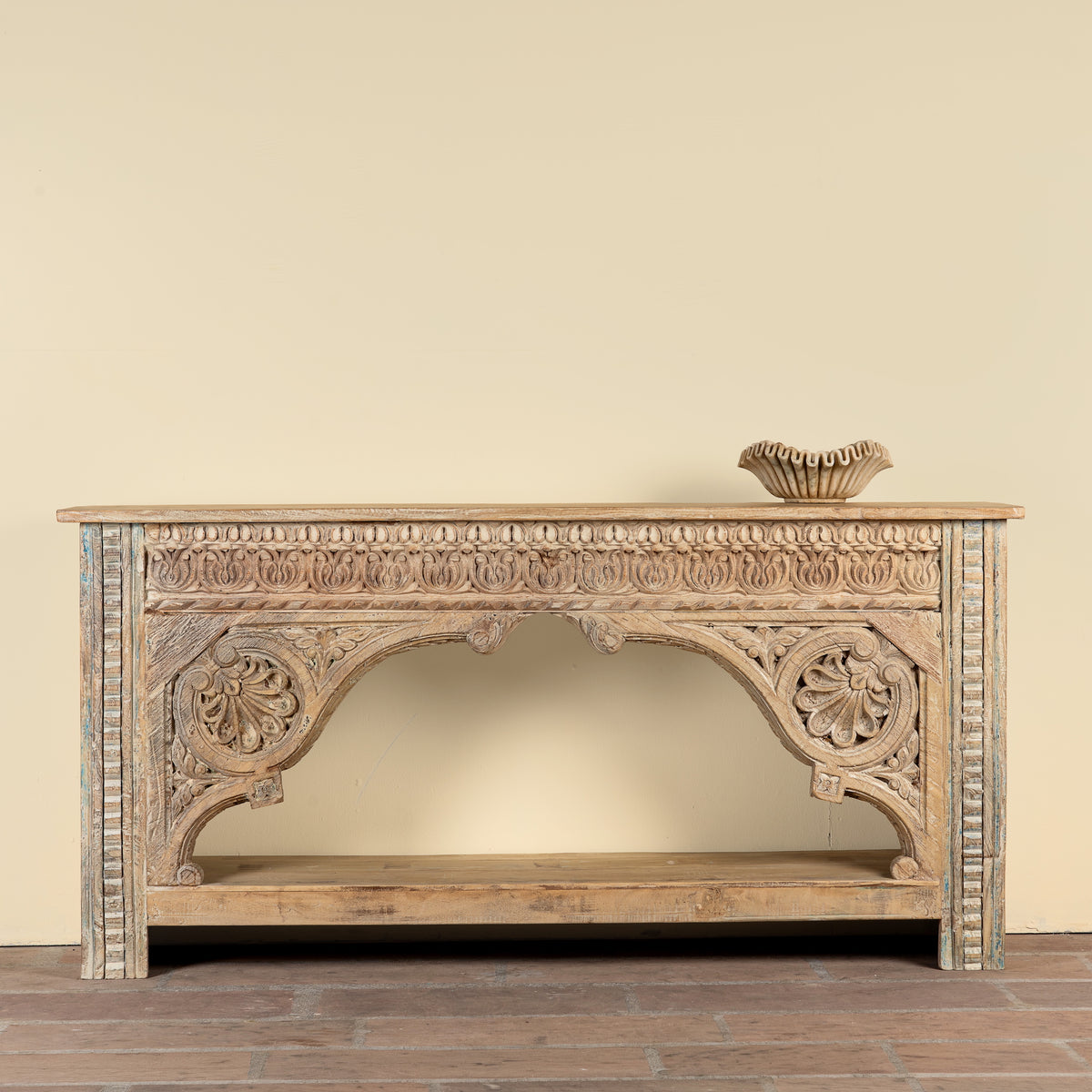 An ornate carved wooden console table with a rustic, weathered finish, featuring intricate floral carvings on the apron and side panels, a lower shelf, and a small decorative bowl placed on top, set against a plain beige wall and dark wood floor.