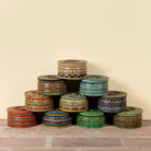 A pyramid arrangement of twelve hand-painted wooden boxes with lids, decorated with intricate traditional patterns in a variety of colors including green, blue, orange, red, and gold, set against a plain beige wall and brick floor.