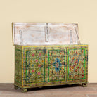 An antique wooden chest with a distressed white-painted lid and an intricately decorated front featuring colorful floral and geometric patterns on a green background, set against a plain beige wall.
