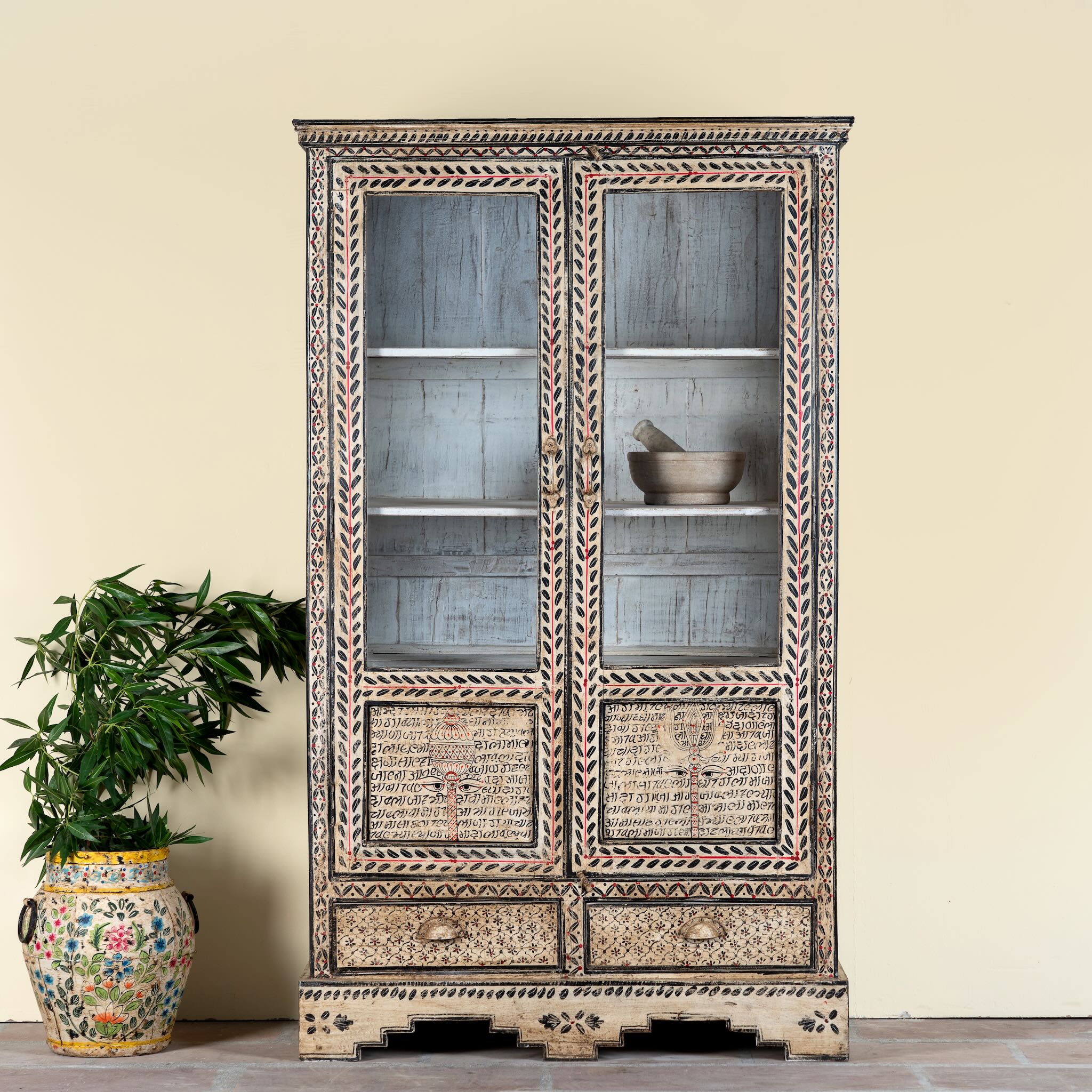 Lifestyle image of a of a hand-painted display cabinet made from recycled wood with glass doors – sustainable furniture featuring Sanskrit motifs and traditional Indian craftsmanship.