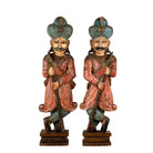 A pair of hand-carved wooden figurines of Indian Rajasthani nobles wearing turquoise and red patterned garments, standing on ornate bases against a white background.