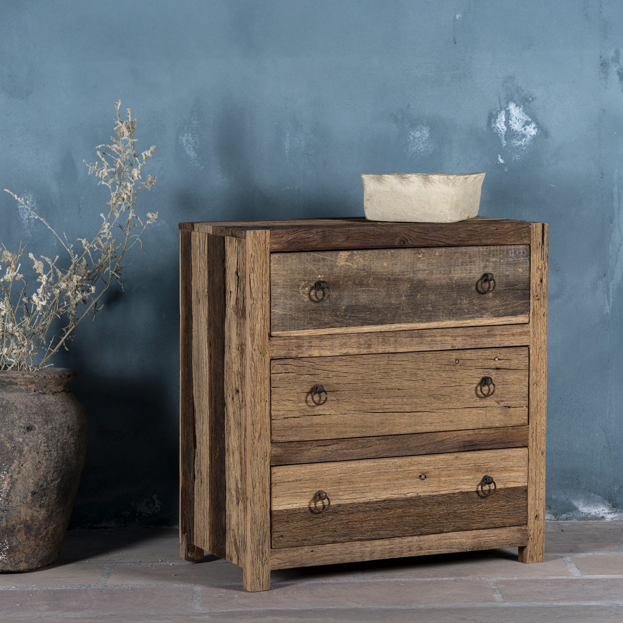 Side view image of a three-drawer dresser in Driftwood/Wabi Sabi style made from recycled old wood, featuring a naturally weathered finish with visible grain patterns and metal ring pull handles for a rustic, handcrafted look.

