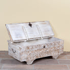 Side angle with open lid of a Shaan mango wood storage chest coffee table with carved front, iron fittings, wooden wheels and cream distressed finish 116cm