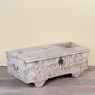 Side angle image of a Shaan mango wood storage chest coffee table with carved front, iron fittings, wooden wheels and cream distressed finish 116cm