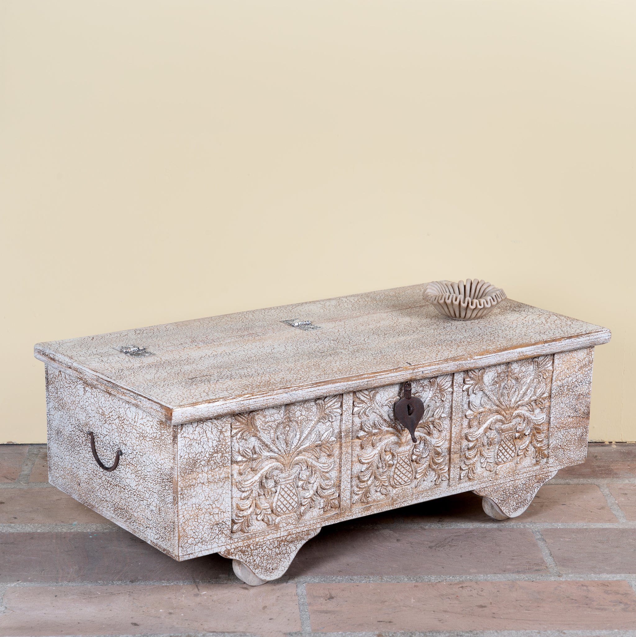 Side angle image of a Shaan mango wood storage chest coffee table with carved front, iron fittings, wooden wheels and cream distressed finish 116cm