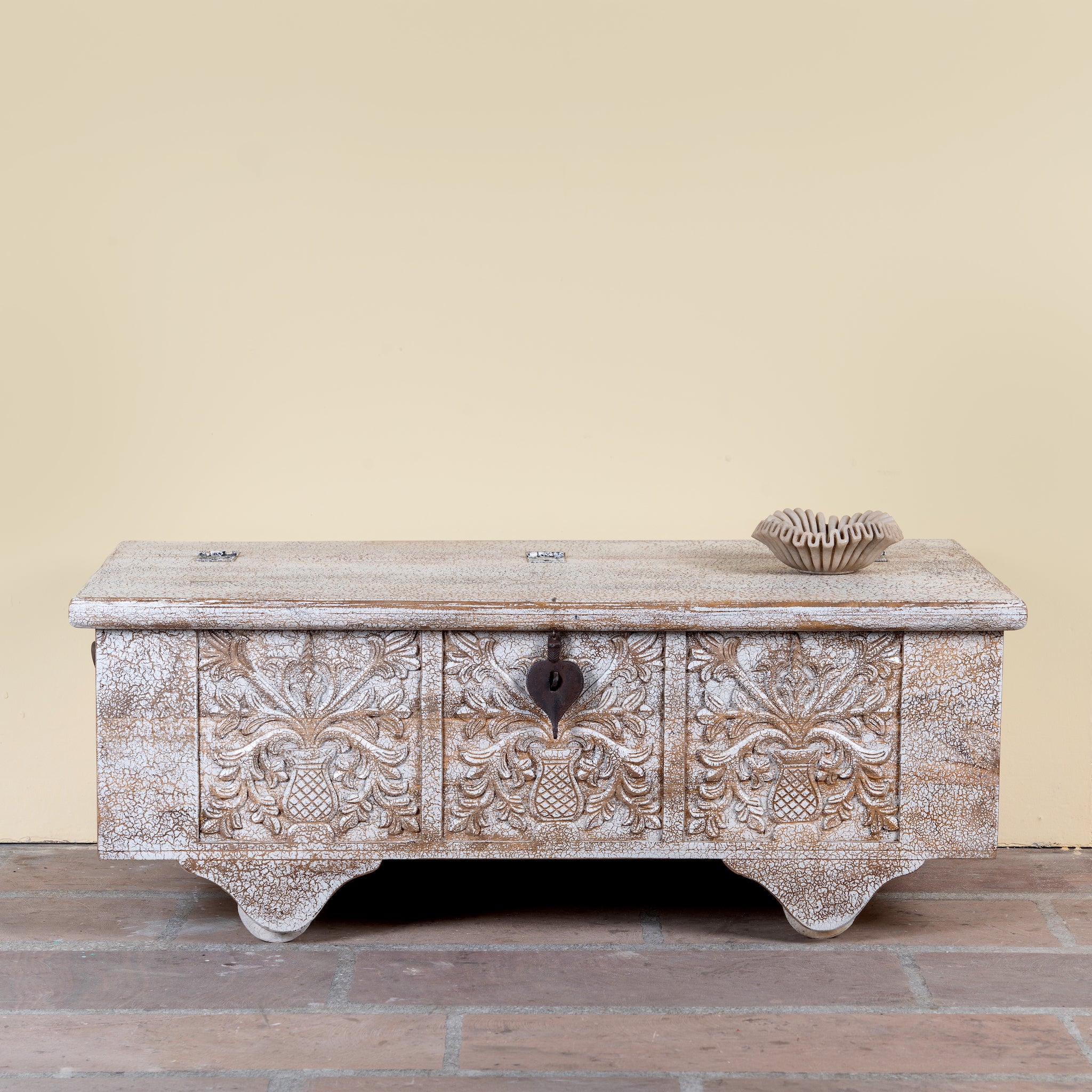 Shaan mango wood storage chest coffee table with carved front, iron fittings, wooden wheels and cream distressed finish 116cm