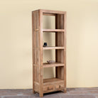 A tall, narrow wooden bookcase with four open shelves and two small drawers at the bottom, shown against a plain beige wall and tiled floor.