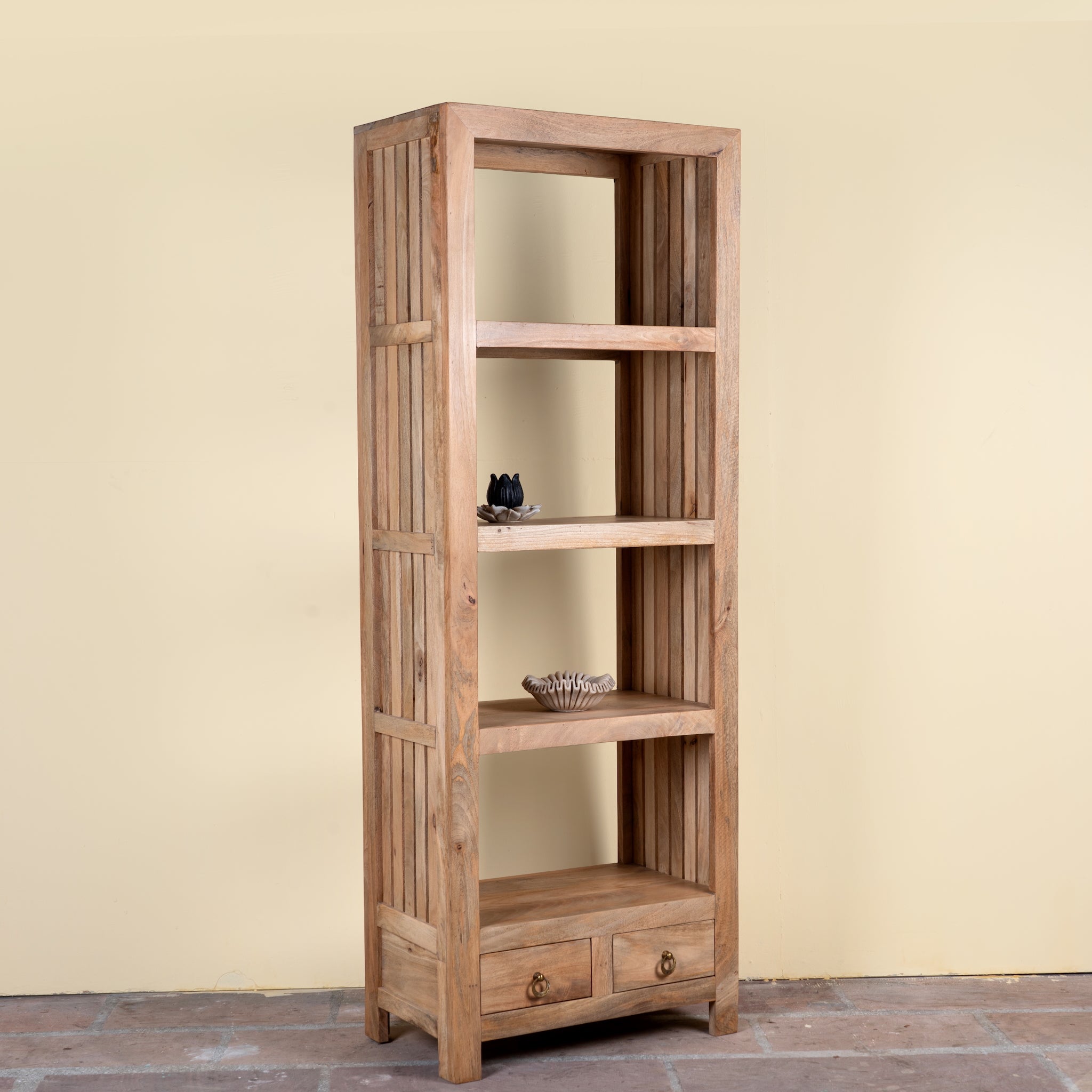 A tall, narrow wooden bookcase with four open shelves and two small drawers at the bottom, shown against a plain beige wall and tiled floor.