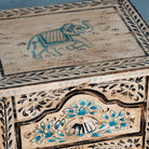 Close up picture of a pair of hand-painted nightstands made from reclaimed wood, featuring floral motifs in soft turquoise and black with intricate carved detailing and two drawers for storage.

