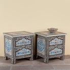 Side angle image of a pair of hand-painted nightstands made from reclaimed wood, featuring floral motifs in soft turquoise and black with intricate carved detailing and two drawers for storage.


